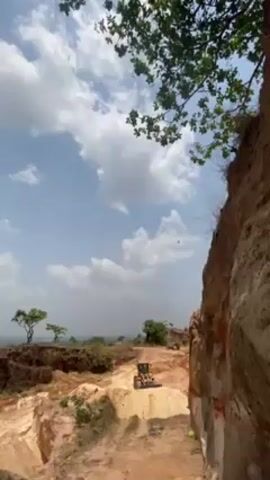 The video shows a view of a construction site in an outdoor area. A bulldozer is visible in the distance, moving on a dirt surface. The surrounding environment features a combination of trees and rocky or sandy cliffs. The sky is partially covered with white clouds. A tree branch with green leaves is visible in the upper portion of the frame, providing a partial foreground to the scene.