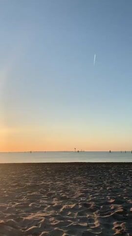 The video opens with a view of a sandy beach and the ocean. The sun is setting on the left side of the frame, casting a warm glow across the sky and water. The sky is a clear blue with a slight gradient towards orange and yellow near the horizon. A faint contrail from an airplane is visible in the upper right corner. The water is calm, with a few distant boats or buoys visible on the horizon. As the video progresses, the sun continues to set, its rays becoming more prominent and creating a reflection on the water. A single, dark object, possibly a beach chair or a lifeguard stand, is visible on the beach in the middle ground. The sand in the foreground shows footprints and texture. The lighting shifts from bright sunlight to a softer twilight glow.
