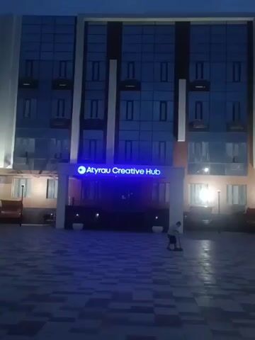 This is a medium shot of the exterior of a modern building at dusk. The building has a glass facade and a sign that reads "Atyrau Creative Hub" illuminated in blue. The ground in front of the building is paved with interlocking stones. In the lower right corner of the frame, a child is visible on a scooter, facing away from the camera. The sky is a dark blue, indicating the end of daylight. There are lights on inside the building, and some outdoor lights are also visible.