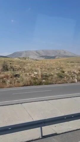 The video shows a view from inside a vehicle, likely a bus, looking out the window at a landscape. In the foreground, there is a road with a white line separating the lanes. To the right of the road, there is a concrete barrier. The background consists of dry, grassy fields with scattered trees and shrubs. In the distance, a large, flat-topped mountain or mesa dominates the horizon under a clear blue sky. A few utility poles and power lines are visible in the middle ground. There are no people visible in the frame, and the only moving object is a white car driving on the road in the middle of the video. The overall impression is a sunny day in a rural, somewhat arid environment.