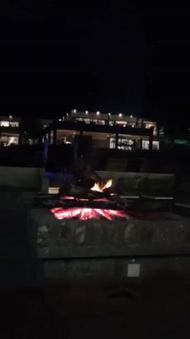 At night, a stone fire pit glows with hot embers and flames, with a blurry hotel building in the background. The sky is dark and stars are visible. The fire pit is surrounded by concrete walls and landscaping. The hotel building has multiple floors and lit windows. There are dark shapes around the fire pit possibly seating or planters. There is chatter, likely people talking, but none can be seen.