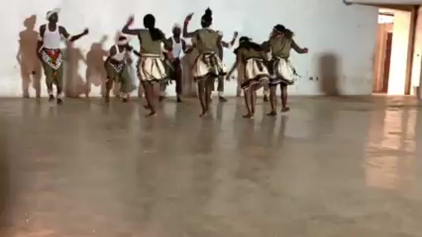 A group of nine people, mainly women, are dancing in a large, sparsely decorated room with smooth concrete floors and white walls. The dancers are dressed in what appears to be traditional attire, including skirts with white and patterned bands, and some wear headbands. The men wear white shirts. They are performing synchronized movements with their arms and legs, appearing to be part of a cultural dance. The room is well lit, casting shadows of the dancers on the wall behind them. A doorway is visible on the right side of the frame, leading to another room. The camera remains still throughout the video, capturing the entire dance performance.