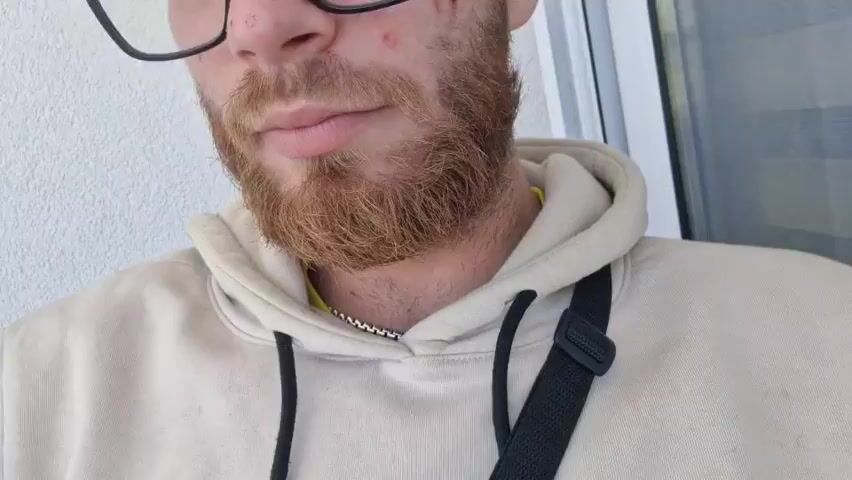 A man with a reddish beard and glasses is smoking a joint. He wears a light-colored hooded sweatshirt and a black strap is visible across his chest. He's in front of a white wall and a window. He takes a puff from the joint and then lowers his hand.