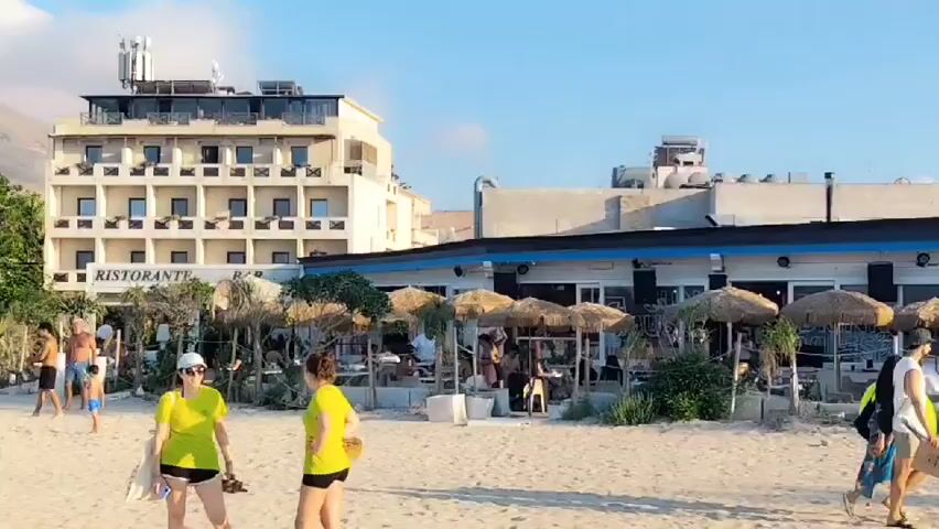 This media file captures a lively scene on a sandy beach, likely in San Vito Lo Capo, Italy, given the prominent rocky mountain formation in the background. The time of day appears to be late afternoon or early evening, indicated by the warm, soft light and long shadows cast across the sand. The weather is clear and pleasant, with a blue sky dotted with fluffy white clouds.

The beach is populated with numerous people enjoying their day. In the foreground, two women walk along the shoreline. One wears a bright yellow t-shirt and a white cap, carrying a bag and what appears to be a surfboard or similar flat object. The other woman is also in yellow and is looking back towards the sea. Further down the beach, a couple walks away from the camera, one of them carrying rolled-up mats or towels.

The scene is lined with beach umbrellas and loungers, suggesting a popular tourist destination. Behind the beach, a multi-story hotel with a prominent "RISTORANTE BAR" sign is visible. Adjacent to the hotel is a building that appears to be a restaurant or beach club, with outdoor seating under thatched umbrellas and patrons seated at tables. Palm trees and lush greenery dot the landscape, adding to the tropical feel.

The overall atmosphere is one of relaxation and enjoyment. People are casually dressed, in swimsuits and summer clothing, and many are engaged in typical beach activities like sunbathing, walking, and socializing. The imposing, rugged mountain in the background provides a dramatic and picturesque backdrop to the bustling beach scene.