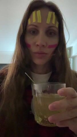 A person with long brown hair is holding a clear glass containing a light yellow liquid with a straw in it. They are stirring the liquid with the straw. They have yellow tape stripes on their forehead and pink markings on their cheeks. They are wearing a brown jacket over a white shirt. The background is a plain white wall.