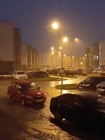 The video shows a nighttime scene during a rainstorm. Streetlights illuminate the scene, casting a yellow glow on the wet pavement and the falling rain. Several cars are parked and driving on a wet street. Buildings line the street, and the overall atmosphere is dark and stormy.