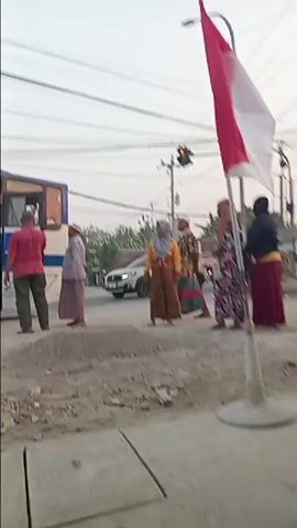 A group of people are standing on a dirt road in front of a blue and white bus. There is a red and white flag in the foreground. The road is littered with debris. Traffic lights are visible in the background. Some of the people are wearing traditional clothing.