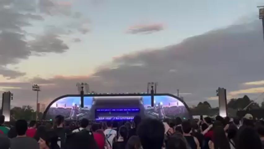 A large crowd of people is gathered at an outdoor concert at dusk. A large stage with screens is visible in the distance. The screens show a performer singing and playing an instrument, possibly a piano or keyboard. The sky is cloudy with a pink hue near the horizon. Lighting towers are on either side of the stage. The crowd is facing the stage and appears to be enjoying the performance.