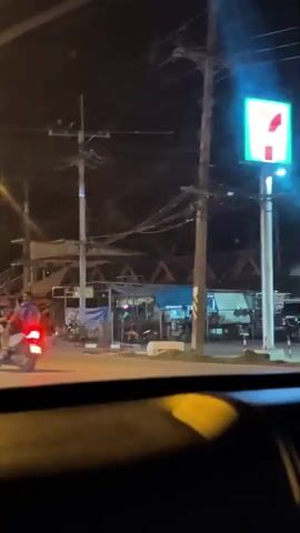 At night, the view from inside a car shows a street with sparse traffic including motorcycles and a small flatbed truck.  The street is lit by streetlights. Overhead are many wires and power poles. There are building structures on the side of the street. The windshield is dirty.
