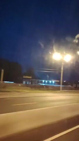 The video shows a view from inside a vehicle traveling on a highway at dusk or night. Streetlights illuminate the scene. On the right side of the road, there are some buildings and trees. The road itself is visible, with lane markings. The sky is dark blue. There are some streaks of light visible, possibly from moisture or dirt on the windshield, or from light sources outside the vehicle. The video is shaky, indicating movement.