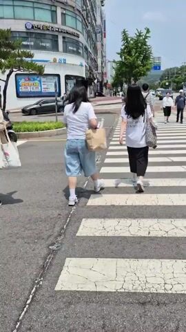 A group of people are crossing a street on a marked crosswalk. The weather appears to be sunny with some clouds. In the foreground, a person wearing a light blue shirt and black pants is walking away from the camera. To the left, two people, one wearing a white t-shirt and denim shorts and the other wearing a white t-shirt and black pants, are also crossing the street. Behind them, several other individuals are visible on the crosswalk, some carrying bags and umbrellas. The background shows buildings lining the street, cars, and trees. The overall environment suggests an urban or suburban setting.