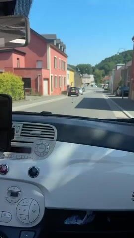 The video shows the interior of a car looking out through the windshield. The car is moving down a street lined with buildings on both sides. The buildings are a mix of styles and colors, with some appearing older and some newer. There are several cars parked along the street and some moving. The street is paved and there is a clear blue sky with no clouds. The overall environment appears to be a small town or village.