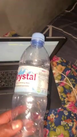 A hand holds a nearly empty bottle of "Brystal" brand water in front of an open black laptop. The bottle is plastic and clear, with a blue cap. The laptop sits on top of a blue floral patterned blanket. The bottle is tilted from vertical towards the laptop. A white charging cable is on the blanket near the laptop.