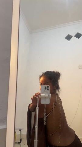 A woman with her hair in a bun stands in front of a mirror in a simple white room, taking a photo of herself with a cell phone mounted on a stand. The woman wears a brown garment with gold chains draped across her bare back. The room has white walls, a white ceiling with two diamond-shaped vents, and a small white stool is visible near the bottom left corner of the mirror. The phone case is white with a decorative charm hanging from it.