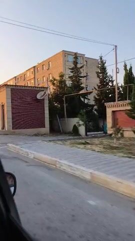 A brief video shows a slow-moving view from a vehicle. The scene includes a building with a light-colored facade, a red brick section, and a satellite dish mounted on the wall. A white truck with a red and white cargo box is parked nearby, and a man wearing a white t-shirt and dark pants is walking away from it. Another building, a multi-story apartment building, is visible in the background, along with trees and a clear sky. The street appears to be paved with asphalt.