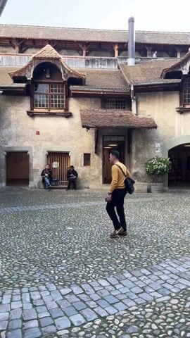The video shows a courtyard with a cobblestone ground and old stone buildings around it. One building has dormer windows and a wooden door that is open. Two people are sitting on a bench in front of the building, and another person is walking across the courtyard. As the camera pans to the right, the viewer sees a small tree in a pot, another building with an open wooden door, and an archway. Two women are standing in the archway, and a young woman is sitting on the ground near the archway, looking at her phone. People are walking through the archway.