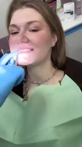 A fair-skinned young woman is sitting in a dental chair. She has braces on her upper teeth and is smiling slightly. A pair of hands wearing blue latex gloves is working on her teeth. The hands are holding a small tool, which is being used to adjust something on her braces. She has a light green dental bib around her neck and a silver beaded necklace with several charms. There are dental tools to the left and a white container on a surface to the right. The lighting is bright, likely from a dental light.