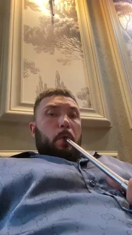 A man with a beard is smoking a hookah. He is wearing a blue shirt with a repeating pattern. The background appears to be a wall with decorative wallpaper. Smoke billows around the man's face and fills the frame.