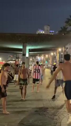 In an outdoor nighttime setting, a group of young men are engaged in a game, apparently of volleyball, although they are using their feet rather than their hands. The players are shirtless or wearing tank tops and appear to be on a wide paved walkway with lampposts and other pedestrians in the background.  There is a covered structure with overhead lighting and pillars nearby. The group is diverse. In the distance is a cityscape with buildings and lights. The overall atmosphere seems relaxed and casual.