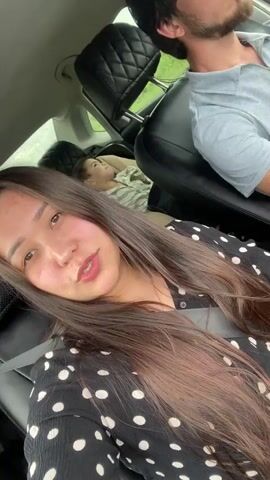 A woman with long, dark hair smiles at the camera while sitting in the passenger seat of a car. She is wearing a black and white polka dot shirt. In the background, a child is asleep in a car seat in the back. A man with dark hair is visible in the driver's seat. The car appears to be in motion, with greenery visible outside the window. The lighting is natural and appears to be daylight.
