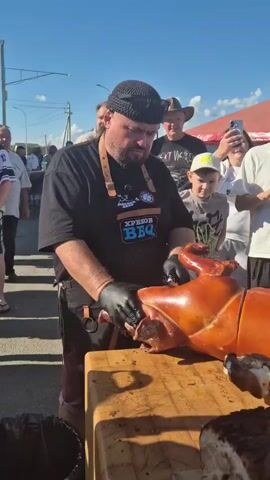 A man wearing a black knit cap, black apron with a logo that reads "ХРЕНоВ BBQ", and black gloves is carving a whole roasted pig on a large wooden cutting board. He is using a large knife to slice off a piece of the pig's shoulder. A crowd of people are watching him, some are taking pictures with their phones. The setting appears to be an outdoor event, possibly a festival or market, with tents and other people in the background. The weather is sunny.