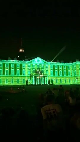 The video captures an outdoor night-time event. A large, light-colored building with many windows is illuminated by vibrant green and yellow lights projected onto its facade. These lights create dynamic patterns that seem to emphasize the building's architectural features, such as windows and columns. In the foreground, a crowd of people is gathered, sitting on the ground and facing the building. Their silhouettes are visible against the illuminated background. The overall atmosphere appears festive and engaging, with the light show serving as the main attraction.