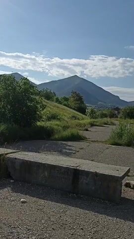 The video captures an outdoor scene, likely in Lermontov, Russia. The time of day appears to be late morning or early afternoon, given the bright sunlight and clear blue sky with scattered white clouds. The weather is pleasant and sunny.

The foreground features a rough, gravelly path with some concrete debris. A large, weathered concrete block serves as a prominent object in the immediate foreground. Beyond this, tall green reeds and grass dominate the landscape, creating a natural, somewhat overgrown feel. In the middle ground, a gentle, grassy slope rises, dotted with more vegetation.

Further in the background, a distinctive mountain, likely Mount Mashuk due to the location context, dominates the horizon. The mountain is covered in green vegetation and its peak is rounded. In the middle distance, between the natural landscape and the mountain, there are signs of human habitation, including houses and buildings.

A key detail is the presence of two people walking through the tall grass in the middle ground. One person is wearing a bright green t-shirt and dark pants, and appears to be walking away from the camera, leading the way. The other person is partially obscured by the reeds but seems to be following. Their activity appears to be a leisurely walk or hike through this natural environment, possibly exploring the area or heading towards the mountain.

The overall mood is serene and natural, with a hint of human presence within the landscape. There is no visible text in the video.