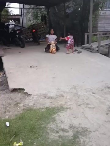 Two young girls are playing on a paved area. The girl in the foreground is sitting on a toy scooter, her legs on either side. She is wearing a white t-shirt and black pants. She has her arms crossed above her head, then she uncrosses them and spreads them out. She is smiling broadly. The other girl is standing behind her, also smiling. She is wearing a pink and white shirt and red shorts. In the background, there is a motorcycle parked under a lean-to structure. There are also wooden structures and some vegetation visible. The ground is a mix of concrete, dirt, and grass.