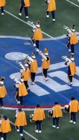 A marching band, seen from a high angle, performs on a football field with a blue and white logo. Band members wear yellow capes, black pants, white shoes, and hats with white plumes. Most carry silver-colored instruments, possibly trombones or baritones, while others carry drums. They move and turn as part of their performance. The field is green with white yard lines.