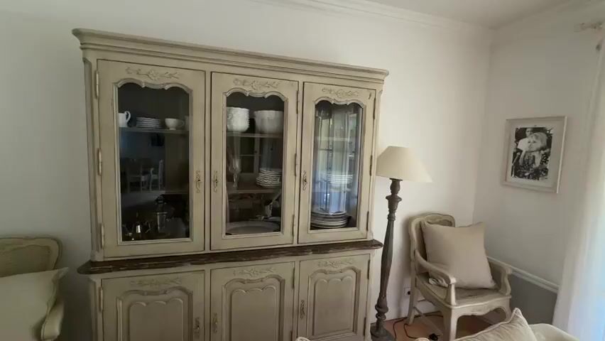The video shows a section of a room with French Country-style decor. 
The room is painted white and has crown molding. On the right side of the initial view is a china cabinet with a dark wood countertop. To the left of the cabinet is a chair with an off-white cushion, and behind the chair is a lamp with a beige lampshade. On the wall is a framed black-and-white photo. Moving towards the left, there is a console table with items on it, including stacks of books and a small container. Then, the camera continues to pan left, revealing a chest of drawers topped by a mirror and several lamps and a fan. To the left of the chest of drawers is an arched doorway, which leads to another room. A white door is located on the wall to the right.
