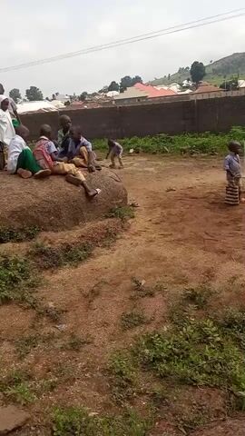 The video shows a group of children in an outdoor, dirt-covered area with some sparse vegetation. In the foreground, a large rock formation is visible with several children sitting on and around it. Some of the children are wearing school uniforms. To the right, a few more children are standing. In the background, there is a wall, some buildings, and a hillside with trees. The sky is overcast. The children appear to be interacting, with some sitting and talking, while others stand and observe. The overall impression is of a school or community gathering in a rural or semi-rural setting.
