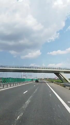 The video shows a view from the perspective of a vehicle driving on a highway. The highway has multiple lanes and is bordered by green guardrails and fields of crops. Several cars and trucks are visible on the road. In the distance, there are tall power lines and towers. A pedestrian bridge crosses over the highway. The sky is partly cloudy with blue sky visible. The video transitions through several shots, showing the vehicle passing under the bridge and encountering other vehicles.