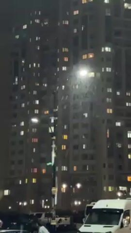 The video shows a nighttime urban scene. A tall apartment building is visible with numerous illuminated windows. In the foreground, there is a street lamp that is lit.  It is snowing, and the snowflakes are illuminated by the streetlight. There are also cars parked along the street. A person in a pink hooded sweatshirt is crouched near a street lamp, partially visible.