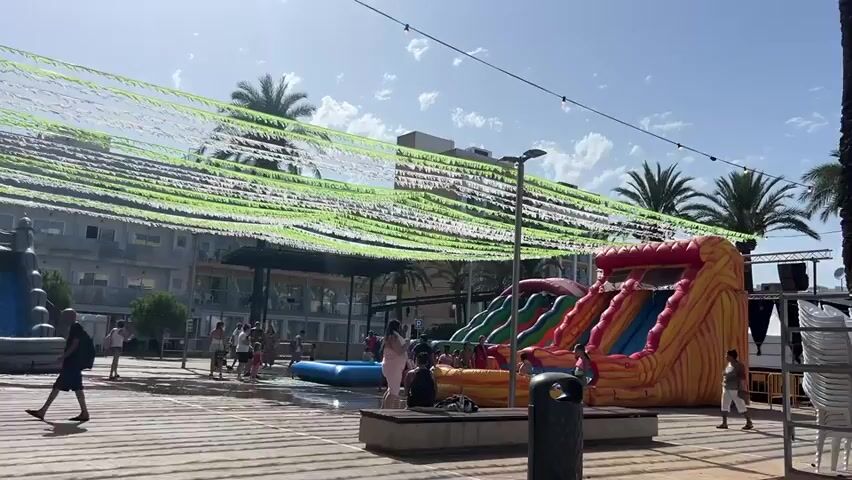 The video shows an outdoor scene with multiple inflatable slides and structures for children. There are people, including adults and children, present in the area. Some children are playing near a blue inflatable pool. The area is decorated with streamers hanging overhead, and there are buildings in the background. The ground appears to be paved and slightly wet in places. In the foreground, a large, colorful inflatable slide dominates the left side of the frame. In the middle, there is a water slide and a smaller inflatable structure. To the right, another large inflatable slide with an orange and yellow design is visible. The overall impression is a festive outdoor event, possibly a children's party or fair.