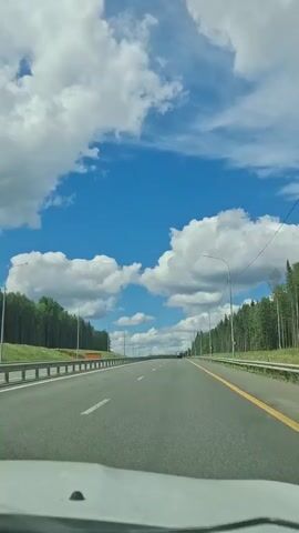The video shows a first-person view from inside a car driving on a highway. The road is paved and appears to be a multi-lane highway with a guardrail on the left and trees lining both sides. The sky is blue with fluffy white clouds. The time stamp indicates the video is about 5 seconds long, from 00:00 to 00:05.