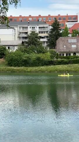 The camera captures a tranquil scene of a river flowing through a residential area. In the foreground, two individuals can be seen paddling a yellow kayak across the water. The river reflects the surroundings, creating a mirrored effect on the surface. Behind the river, a lush green bank is lined with vegetation. Beyond this greenery, a collection of houses and apartment buildings of varying styles rises in the background. These buildings are painted in a range of colors, from white to orange-red, contributing to the neighborhood's diverse appearance. As the camera pans to the right, a footbridge comes into view, spanning the river. The bridge, constructed with a metal framework, offers a walkway for pedestrians. The sky above is partly cloudy, casting a soft light over the entire scene.