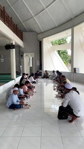 A group of people, predominantly men and boys, are sitting on the floor of a large, open-air building, likely a mosque or community hall, sharing a meal. They are arranged in two long rows facing each other, with individual meal boxes placed in front of each person. The floor is tiled, and the ceiling is exposed with structural beams. Sunlight streams in from an open side of the building, illuminating the scene. The people are dressed casually, with some wearing traditional Islamic attire like kufi caps. Some are actively eating, while others appear to be conversing. In the background, there are religious artifacts like green prayer mats and a speaker system. The overall atmosphere seems communal and peaceful.