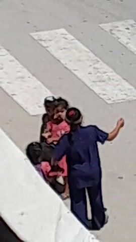 An aerial view shows four young children and an adult on a crosswalk. The adult, wearing a dark blue outfit and a visor, stands with their back to the camera. Three children are in front of the adult. One child, on the far left, wears a patterned shirt and shorts and a dark cap. Another child, in the center, wears a pink dress. The third child, on the far right, also appears to be wearing a pink dress. A fourth child, partially obscured, is kneeling in front of the other three, wearing a pink and purple striped garment and a backpack. The children are interacting with the adult and each other. The ground is a paved surface with white crosswalk stripes.