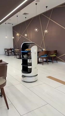 A food delivery robot is stationary in the middle of a restaurant aisle. The robot is white and tall with multiple shelves for carrying food. The top shelf has a plate of food on it. The robot has a digital display on its side that shows the number 12. The restaurant has brown and white tiled floors and brown walls decorated with geometric patterns. There are tables and chairs around the robot. The lighting is dim, with pendant lights hanging from the ceiling.