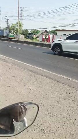 The video appears to be shot from the perspective of someone riding a motorcycle on a paved road. In the foreground, a motorcycle's side mirror is visible, reflecting a portion of the rider. The road itself is grey asphalt with white lane markings. On the right side of the road, there is a grey gravel shoulder. Beyond the road, there are various structures including buildings with white walls and red accents, a green gate, and trees. Power lines and tall utility poles are visible against a bright, hazy sky. Several vehicles are seen moving on the road, including a white SUV, a blue and white bus, and multiple motorcycles. The overall environment suggests a somewhat rural or suburban roadside setting during daylight.
