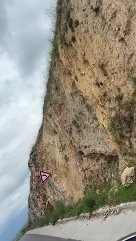 The video shows a road winding along a steep hillside on a cloudy day. The camera is positioned from a vehicle moving along the road. The hillside to the right is rocky with sparse green vegetation and a few red flowers. A road sign warning of a sharp left turn is visible near the beginning of the video. The slope of the hill gradually transitions from rocky to grassy further along the road. Power lines can be seen crossing the hillside. The sky is filled with gray clouds.