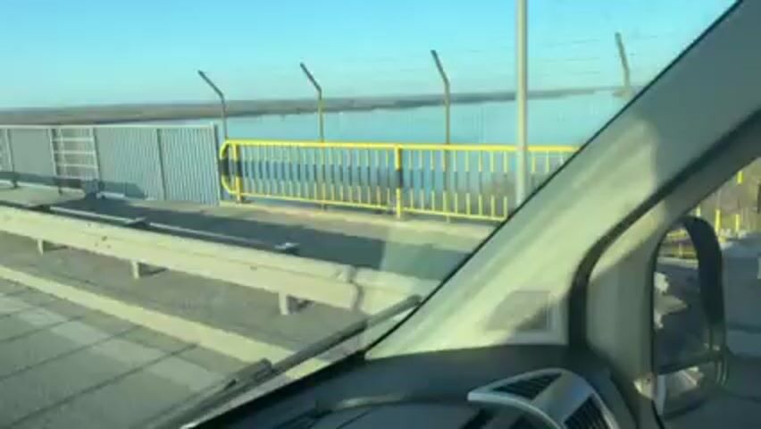 The video shows the view from the passenger seat of a vehicle crossing a bridge. The water is visible outside the passenger window, with a flat, green shoreline in the distance. The bridge is gray and has a metal railing. The sky is clear and blue.