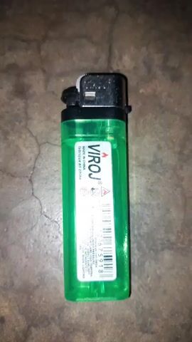 The video shows a green Viroj lighter lying on a cracked, dirty floor. The camera focuses on the top of the lighter, showing the spark wheel and gas nozzle.