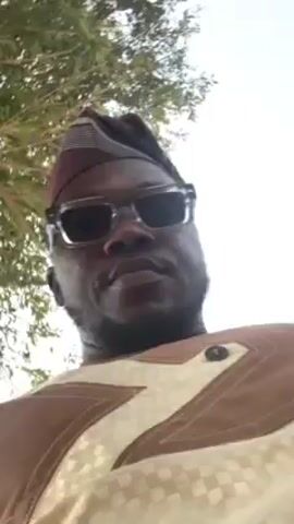 A man, seen from the chest up, looks into the camera. He is wearing a traditional patterned hat and tinted glasses. He is also wearing a tan and brown shirt with patterned details. He is outside under a tree. The sky is visible.