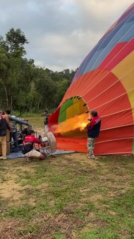 A group of people are gathered around a large, colorful hot air balloon that is partially inflated on a grassy field. A burner is being used to heat the air inside the balloon, creating a large flame that is visible through the opening of the balloon. One person is operating the burner, while others are holding onto the balloon or the burner equipment. The balloon is colorful, with stripes of red, orange, yellow, green, blue, and purple. The background shows trees and a cloudy sky. The scene appears to be an outdoor event or activity, possibly a hot air balloon launch.