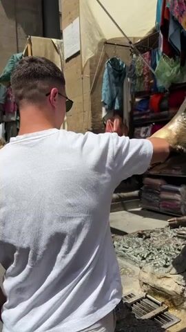 A young man in sunglasses and a white long-sleeved shirt touches the snout of a bronze boar statue. The man smiles and steps away from the statue, which is near a fountain. A vendor selling scarves and other textiles has a stall nearby. The scene appears to be outdoors on a sunny day.