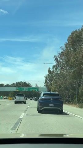 The video shows a highway scene from the perspective of a driver. In the foreground, the dashboard and windshield frame the view. Ahead, a blue Volkswagen ID.4 electric SUV is driving in the right lane. To the left of the ID.4, other vehicles are visible, including a white SUV and a dark sedan. Above the highway, a green overhead sign indicates directions for "SOUTH 280 San Jose EXIT 58" and "NORTH 280 San Francisco EXIT 5A". Trees and foliage line the right side of the highway, and the sky is blue with scattered white clouds. The road appears to be in good condition, and the traffic seems moderate. The overall impression is a typical driving scene on a sunny day.