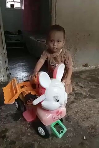 A young child, wearing a brown t-shirt and shorts, is kneeling on a dirty floor in front of a toy truck with a rabbit-shaped attachment. The child is holding onto the toy and looking towards the camera. The background appears to be an interior space with a doorway and a wall. The lighting is dim.