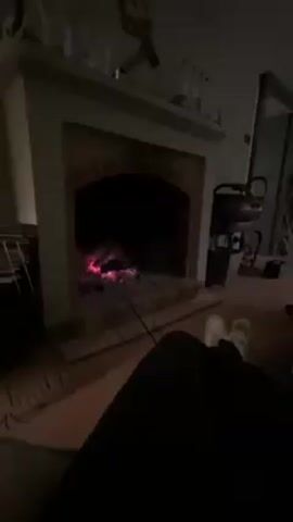 The video pans from a person's feet resting in front of a fireplace to a man sitting on a couch, using his phone. The fireplace is brick and has a green mantle with objects on it. The man on the couch is wearing a dark shirt and a green beanie. A coffee table in front of him has books, magazines, bottles, and a glass of amber liquid on it. There are more rooms visible in the background, including a partially open door. Music is playing in the background.