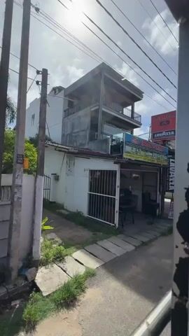 The video, shot from a moving vehicle, captures a street scene in Talpe, Sri Lanka, during the daytime under sunny skies with some clouds.

The initial view shows "CAFE AROMA" and "SL IRON SHOP" signs. The "SL IRON SHOP" sign advertises "Kitchen and Cafe Equipment, Interior & Exterior Decorating, Landscape Garden, Furniture for Interior Decorating, Landscaping," with an address "Unawatuna, Galle" and a phone number "077 578 5535." Both shops display "LAVAZZA ITALY'S FAVOURITE COFFEE" branding.

As the vehicle moves, a "GROCERY" store with "Gamini STORES" signage and "Fortune" and "Edburough" brand advertisements becomes visible. A red tuk-tuk is parked near the grocery store. Electrical poles with numerous overhead wires are prominent throughout the scene.

Further along, a modern building housing "CRYSTAL CONSTRUCTION" is seen, with two white cars parked outside. Another sign for "CRYSTAL CONSTRUCTION CONSORTIUM" lists "DESIGN," "CONSTRUCTION," and "SALES." The street appears to be paved in some sections and unpaved/dirt in others, with grass and bushes growing along the side.